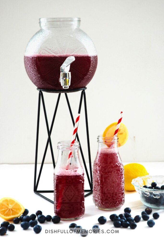 Drink dispenser on tall stand filled with blueberry lemonade. Two small bottles in front and lemons and blueberries next to them on the table.