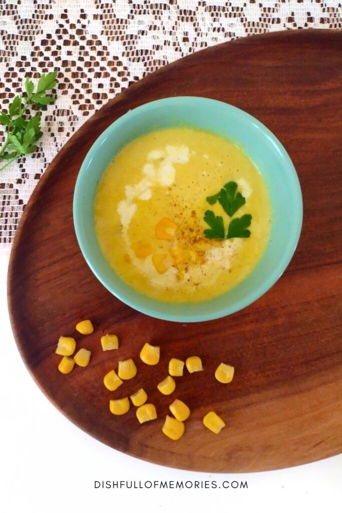 Bowl of yellow creamy corn soup in a bowl on a wooden serving tray with corn kernels around it.