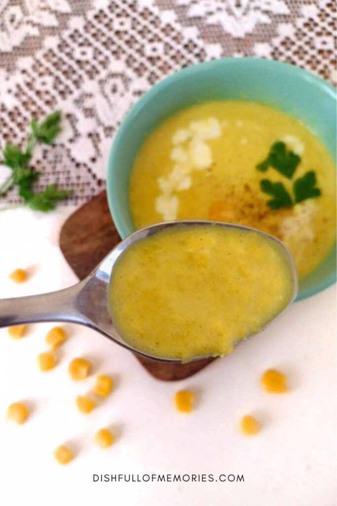 Close up of a spoon filled with yellow corn soup. Bowl and corn kernels in the background.