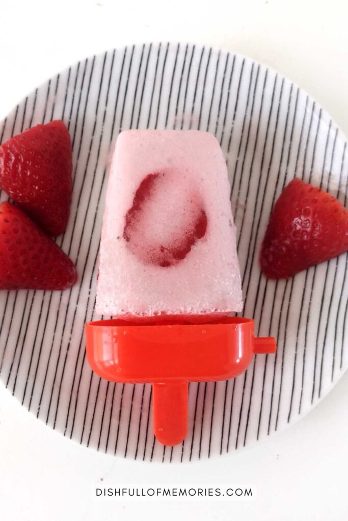 Strawberry popsicle on red base displayed on striped plate with fresh strawberries next to it.