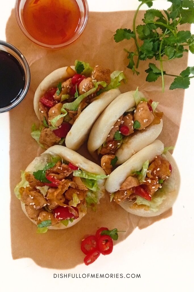 Four homemade asian bao buns filled with chicken and vegetables on a piece of parchment paper. Cilantro, chili pepper and dipping sauces around them.